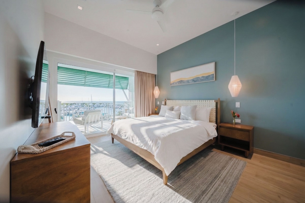 A modern bedroom with a large bed, a flat-screen TV, a desk, and a view of the outdoors.