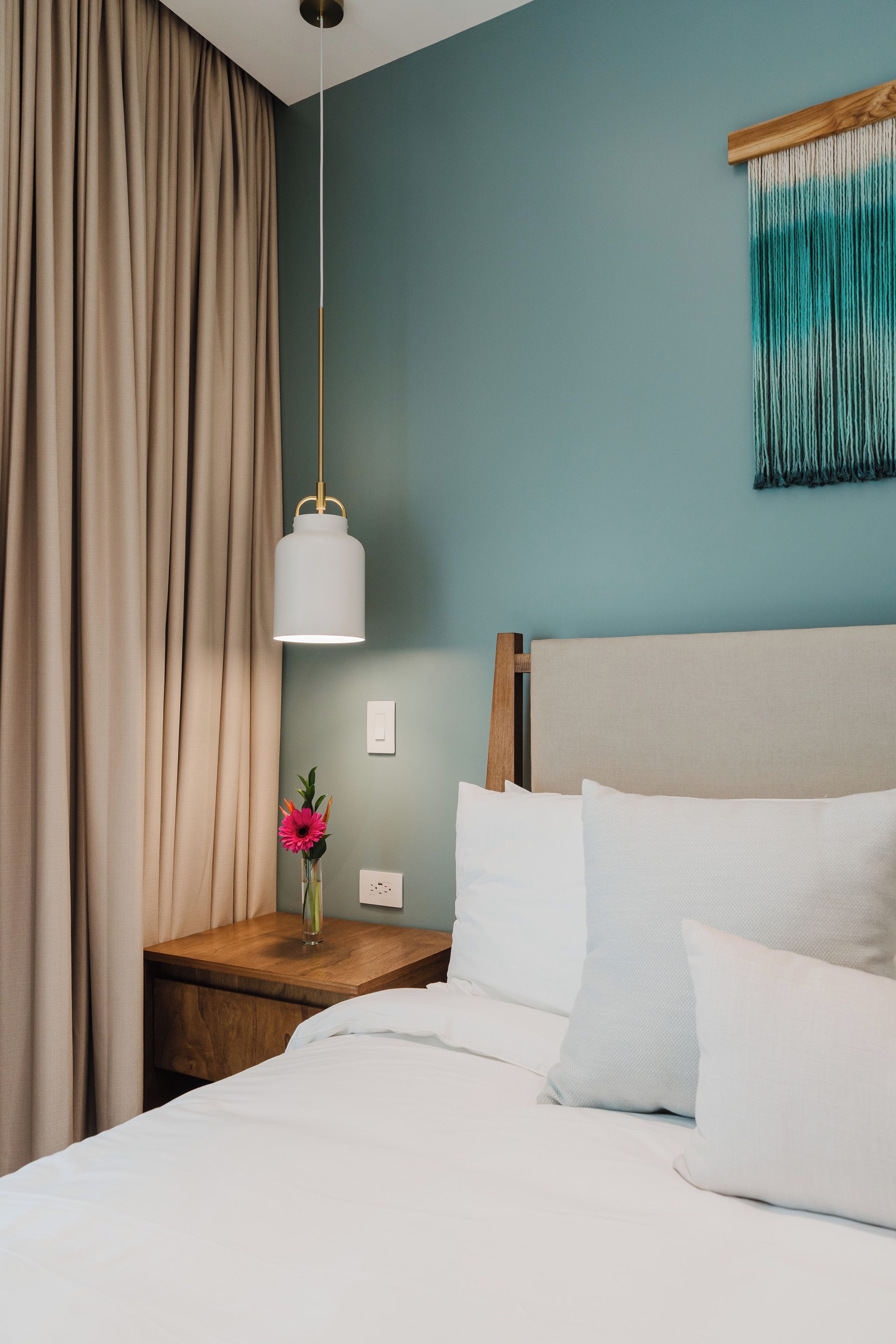 A hotel room with a bed, a nightstand, a vase with a flower, a hanging light fixture, and a wall-mounted tapestry.
