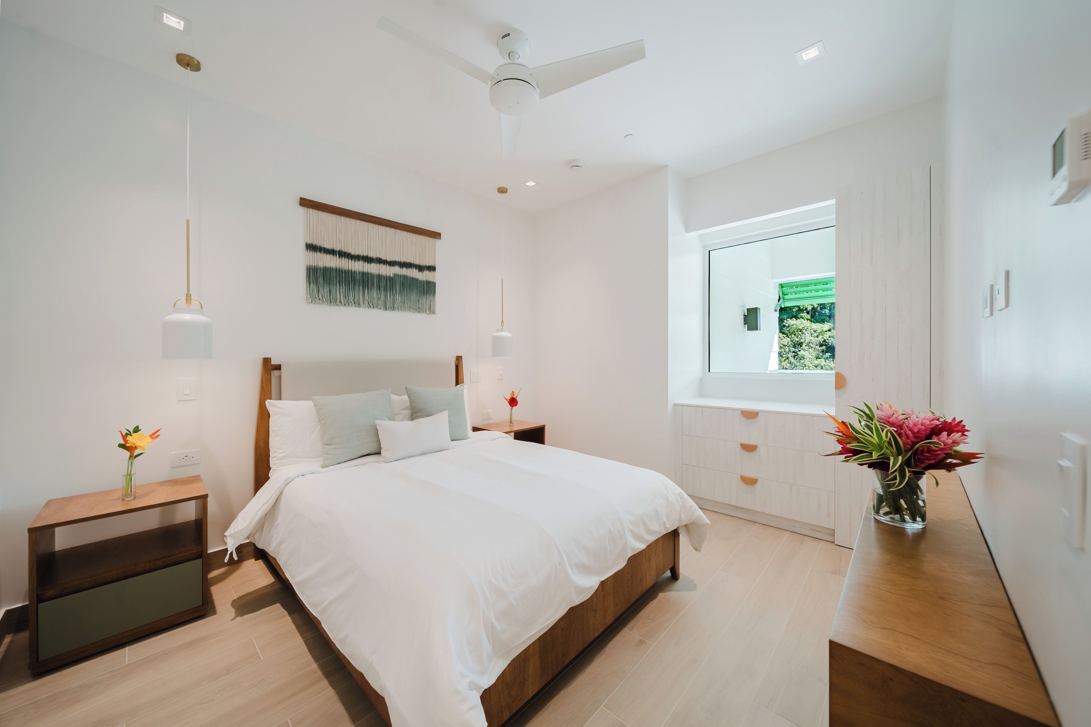 Une chambre moderne comprenant un lit, des tables de chevet, un ventilateur de plafond, une œuvre d’art accrochée au mur et une fenêtre offrant une vue agréable.