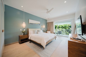 A bedroom with a large bed, a nightstand, a desk, a television, and a view of a balcony with greenery. - Las Villas Marina Pez Vela  (Provincia de Puntarenas)
