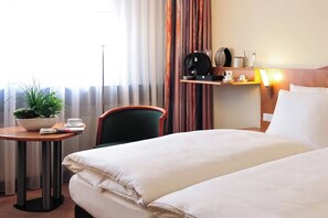 Ein Hotelzimmer mit einem Bett, einem kleinen Tisch mit Pflanze, einem Sessel, einem Regal mit Kaffeemaschine und einer Lampe.