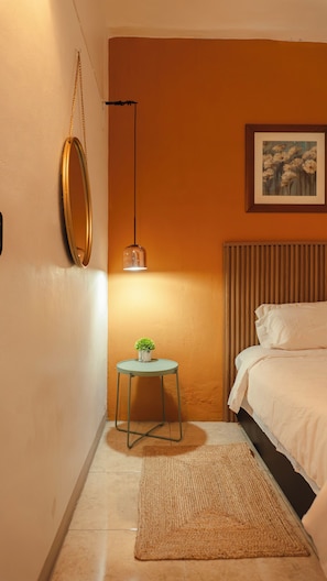 Ein Schlafzimmer mit einem Bett, einem Nachttisch mit Pflanze, einem hängenden Spiegel, einem gerahmten Bild an der Wand und einer Stehlampe.