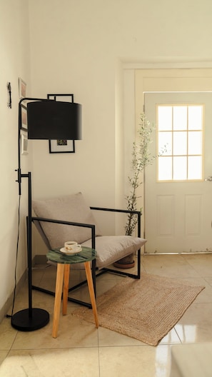 Ein Zimmer mit einem Sessel, einem kleinen Tisch mit einer Tasse, einer Stehlampe, einer Pflanze und einer Tür mit einem Fenster.