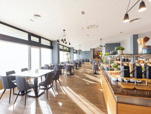 Een ruime eethoek met meerdere tafels en stoelen, een buffet met diverse gerechten, en grote ramen voor natuurlijk licht.