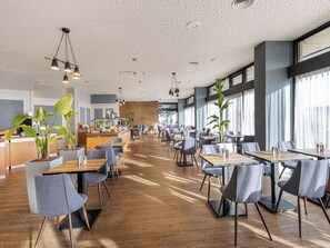 Een ruim restaurant met houten tafels en stoelen, grote ramen en binnenshuis planten.