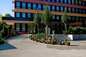 Een modern gebouw met een meerlaags structuur, een overdekte ingang en een aangelegde omgeving met bomen en bloemen.