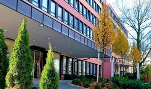 Een modern gebouw met een glazen gevel, een overdekte ingang en een aangelegde tuin met naaldbomen.