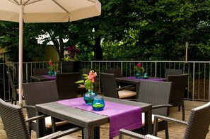 Een terras buiten met rieten meubels en paarse tafelkleden, omringd door een metalen hekwerk en weelderige begroeiing.