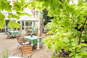 Un patio al aire libre con mesas y sillas, rodeado de follaje y edificios al fondo.