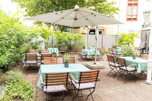 En udendørs terrasse med borde og stole under parasoller, omgivet af grøn vegetation.