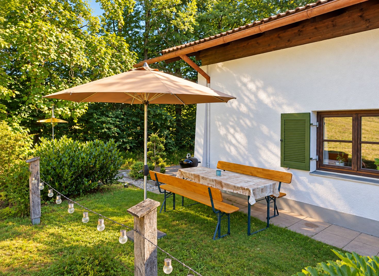 Eine Terrasse mit Tisch und Bänken, umgeben von Grünpflanzen und einem weißen Gebäude mit einem Fenster.