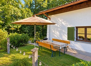 Eine Terrasse mit Tisch und Bänken, umgeben von Grünpflanzen und einem weißen Gebäude mit einem Fenster.