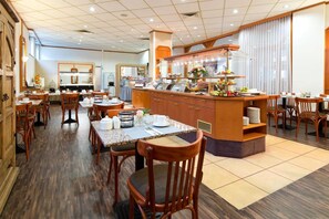 Un comedor de hotel con mesas y sillas de madera, una barra central con exhibición de frutas y un ventanal con cortinas.