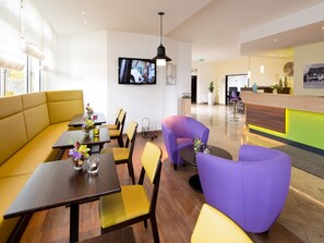 Una hall d'hotel moderna con sedute gialle e viola, una reception e un televisore a schermo piatto montato sulla parete.
