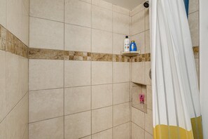 A shower area with beige tiles, a shower curtain, and a shelf with toiletries. - Amazing 5-bedroom house with AC in charming MD minutes from DC and VA (Fort Washington)