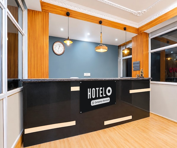 A hotel reception desk with a sign indicating it is company-serviced. There are two clocks on the wall, a window with a view, and pendant lighting.