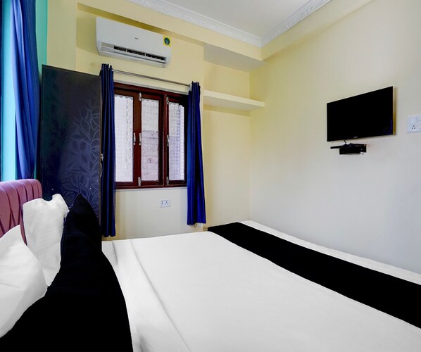 A hotel room with a bed, a television, an air conditioning unit, a window with curtains, and a wall-mounted TV.
