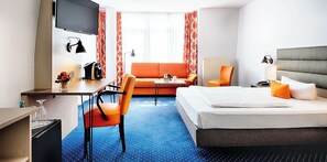 Ein Hotelzimmer mit Bett, Schreibtisch, Sessel, Sofa und Fernseher.