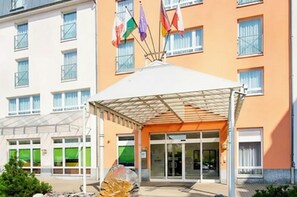 Ingresso di un hotel con una tettoia, diverse bandiere e un ingresso con porta in vetro.