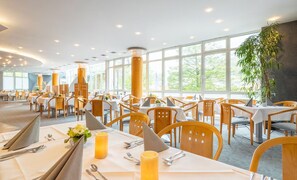 Ein großzügiger Essbereich mit mehreren Tischen und Stühlen, großen Fenstern und einer zentralen Säule.