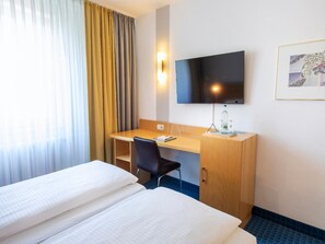 Ein Hotelzimmer mit zwei Betten, einem Schreibtisch, einem Sessel, einem Fernseher und einem gerahmten Bild an der Wand.