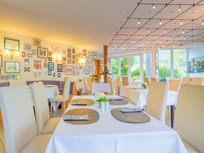 Ein Restaurant mit weißen Tischdecken, beigen Stühlen und gerahmten Bildern an den Wänden.