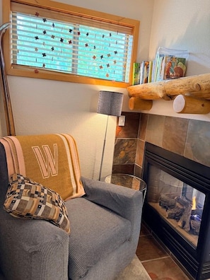 Una sala de estar acogedora con chimenea, un sillón cómodo, una ventana con persianas y una estantería con objetos decorativos.