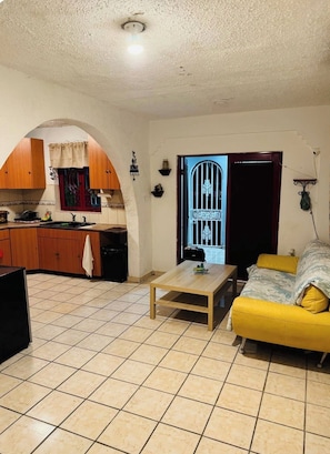 A living room with a yellow sofa, a wooden coffee table, and a kitchen area with wooden cabinets and a tiled floor. - Huesca House next to Airport (San Juan)