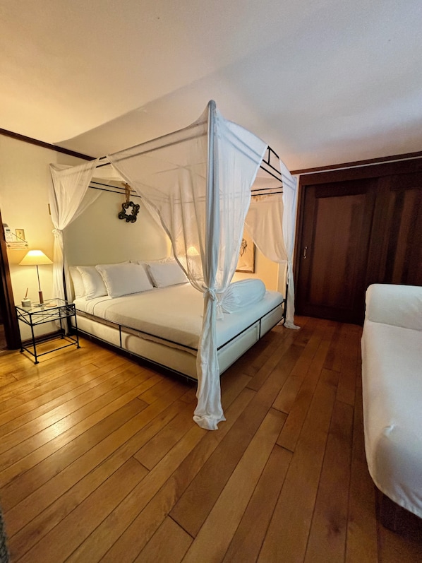 A bedroom with a four-poster bed, a bedside table, a lamp, and a wooden floor.