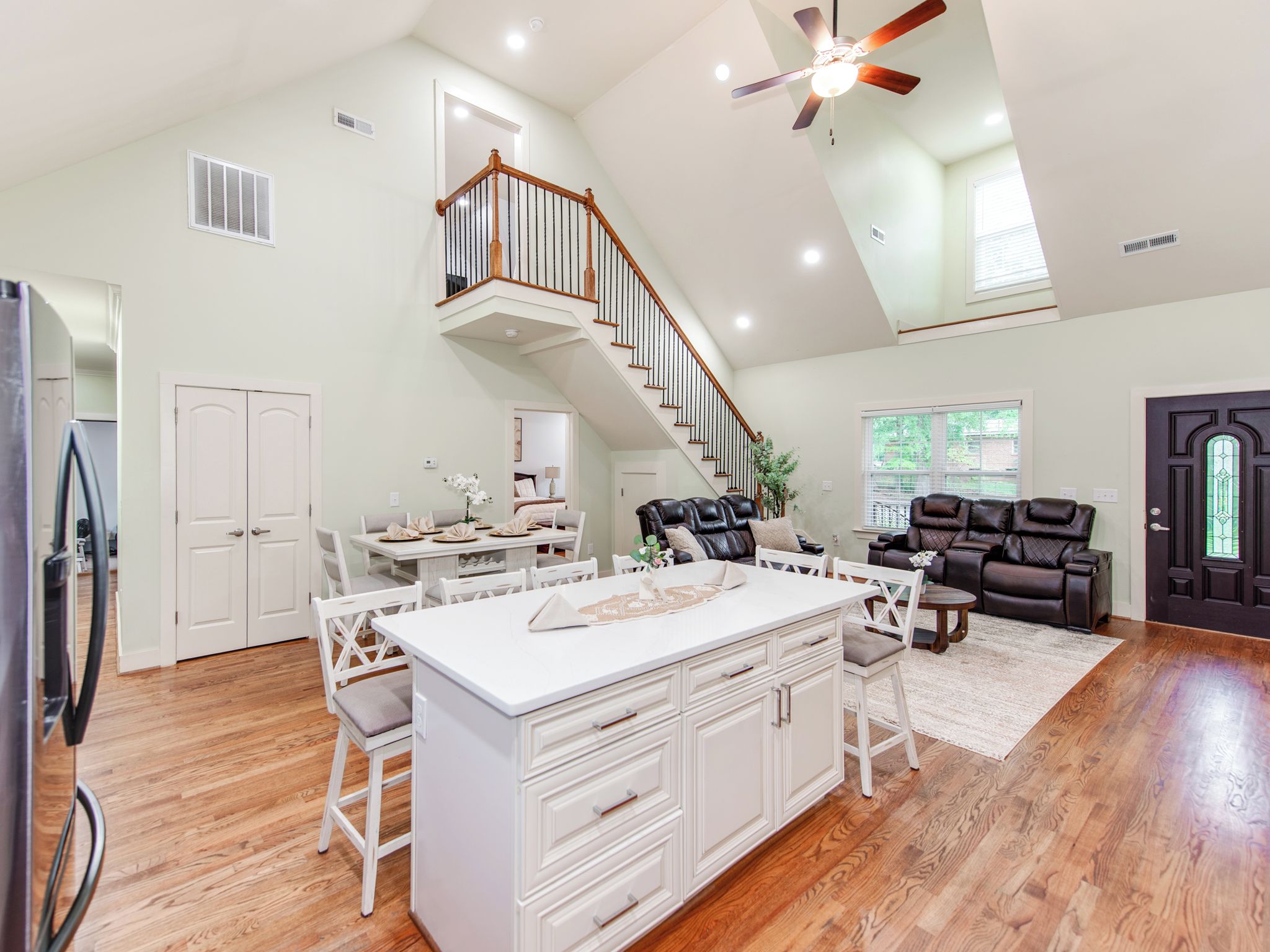 A spacious kitchen with a central island, a dining area, a living room with a leather sofa, and a staircase leading to an upper level.