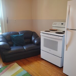 A compact kitchen with a white stove, oven, and refrigerator. A black leather sofa with a teal pillow is positioned against the wall.
