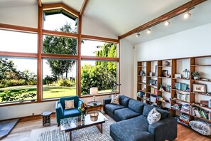 A spacious living room with large windows, a bookshelf, and a coffee table. - Water & Gulf Islands Views, Beach Access, Hot Tub, Game Room (Bellingham)