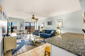 A modern living room with a blue sofa, a dining table, and a kitchen area with granite countertops. - Sunrise Symphony  pool, ocean views, and beach access (Duck)