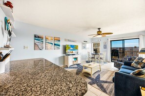 A modern living room with a kitchen area, a dining set, and a large window offering a view. - Sunrise Symphony  pool, ocean views, and beach access (Duck)
