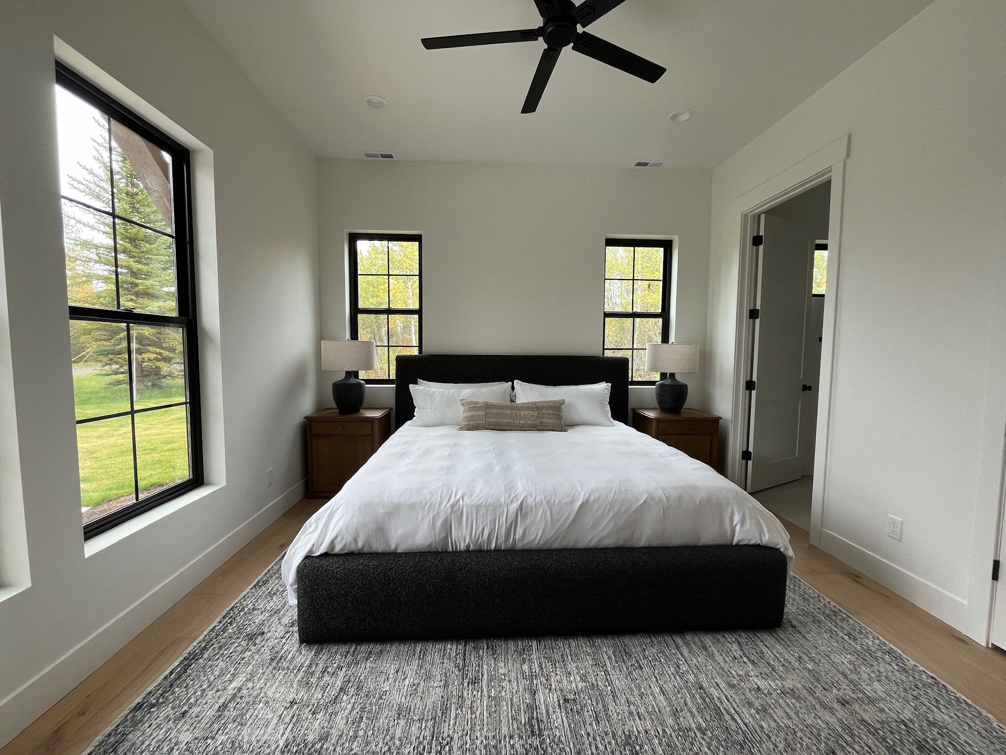 A bedroom with a large bed, two bedside tables, a ceiling fan, and windows with a view of trees.