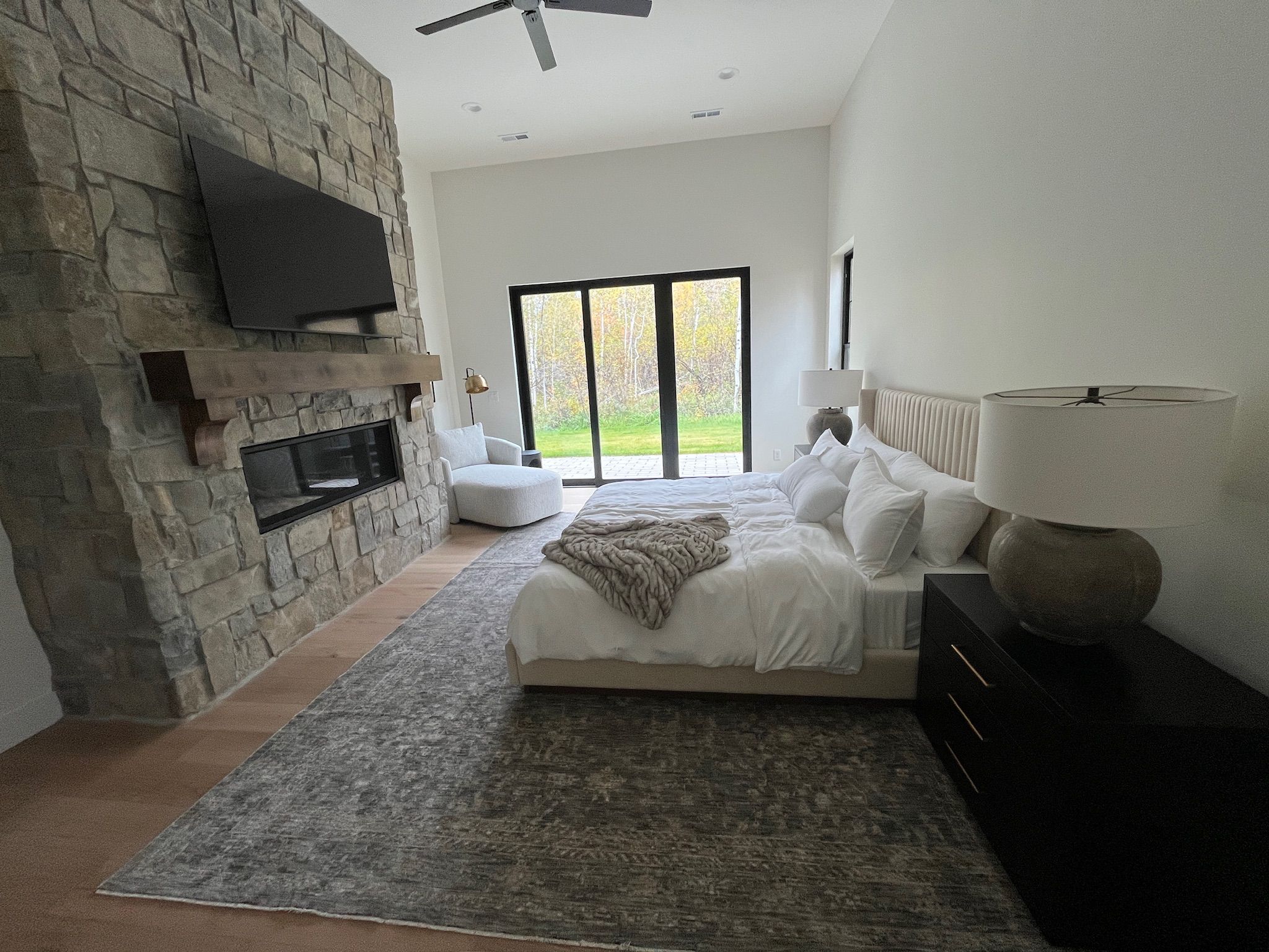 Een moderne slaapkamer met een stenen openhaard, een groot bed met wit beddengoed, uitzicht op het landschap door een schuifdeur en een plafondventilator.