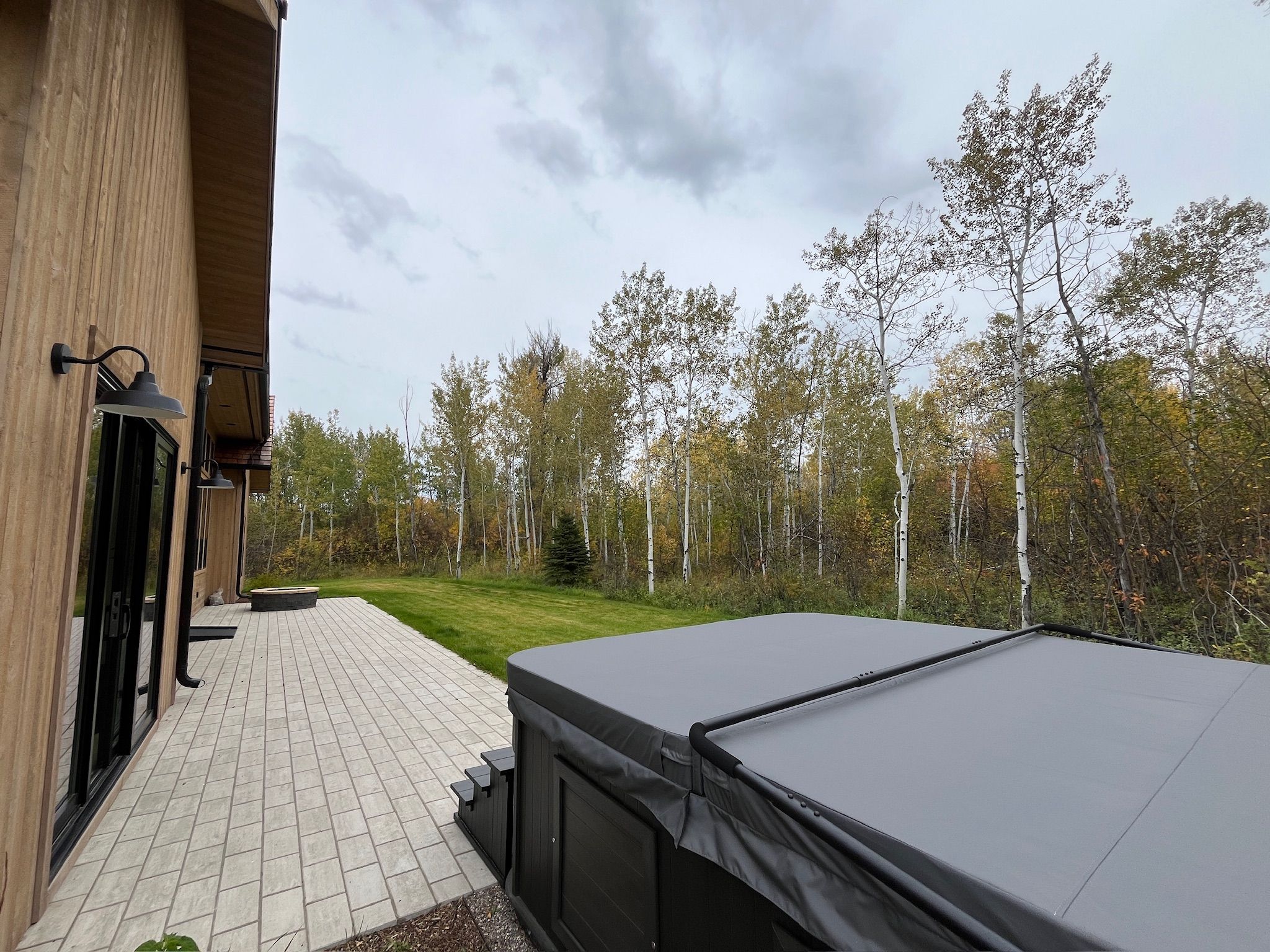 A patio with a covered hot tub, a brick pathway, and a wooden building with a window and a wall-mounted light fixture.