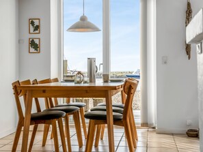 Un espace repas avec une table et des chaises en bois, un luminaire suspendu et une vue sur la mer grâce à de grandes fenêtres.