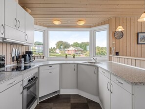 A modern kitchen with white cabinets, a built-in oven, and a large window offering a view of a house and greenery. - "Taneli" - 400m from the sea by Interhome (Glesborg)