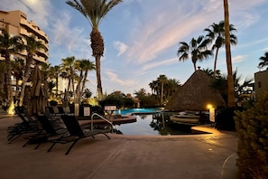 Ein Poolbereich eines Resorts mit Liegestühlen, einer Reetdachkonstruktion und Palmen.