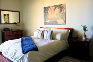 Ein Schlafzimmer mit einem Bett, einem Schminktisch, einem Nachttisch, einer Pflanze im Blumenvasen und einem Bild an der Wand.