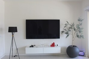 Ein modernes Wohnzimmer mit einem großen Flachbildfernseher an der Wand, einem weißen Konsolentisch mit Dekoration, einer Stehlampe und einem Blumentopf.