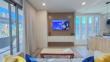 Una sala de estar moderna con un televisor de pantalla plana grande montado en una pared de madera, un sofá con cojines estampados y vistas al exterior a través de amplios ventanales.