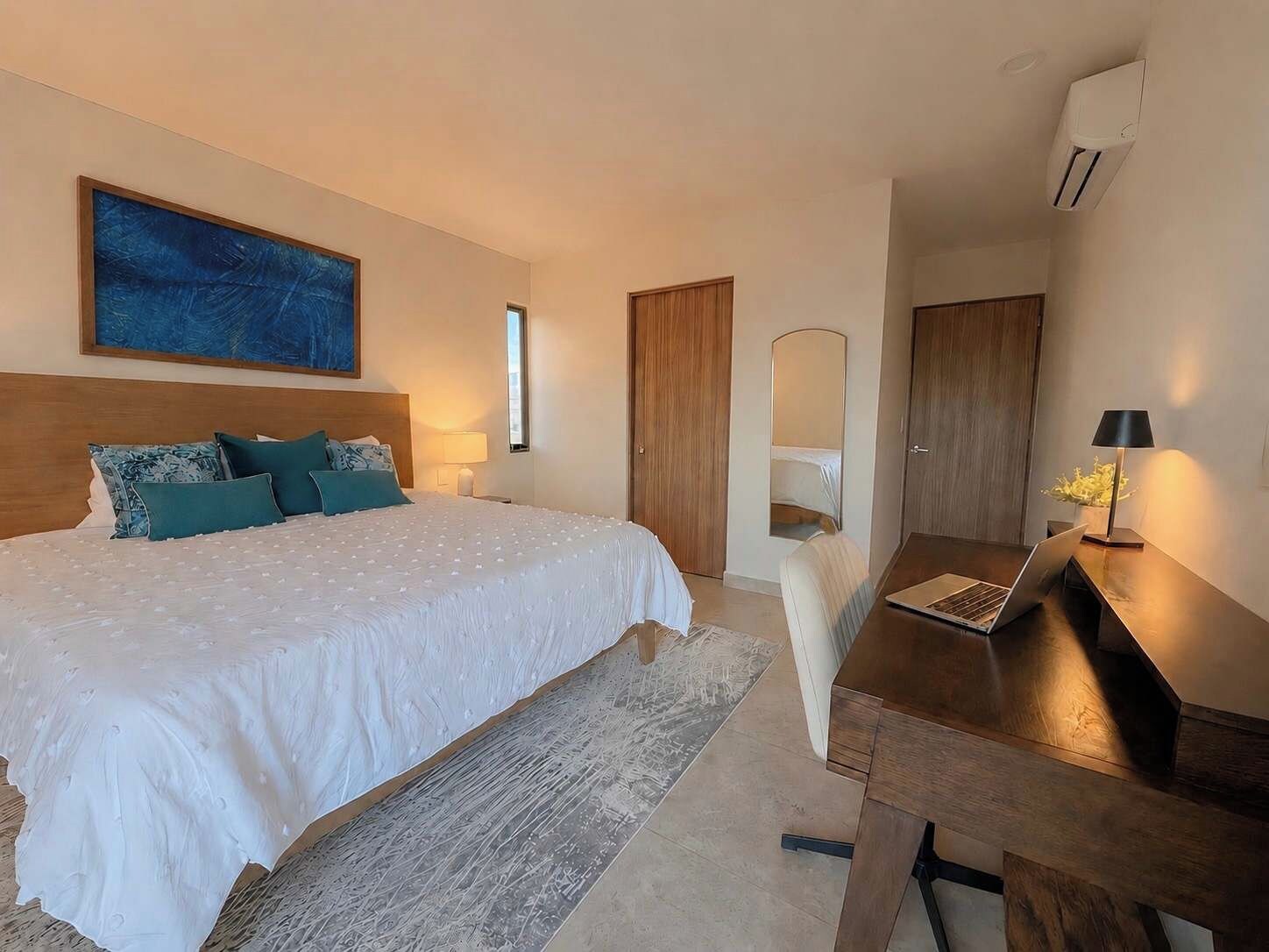 Une chambre à coucher avec un lit, un bureau avec un ordinateur portable, un miroir et un tableau accroché au mur.