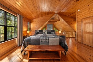 A cozy attic bedroom with a wooden sloped ceiling, a large bed with a quilt, a bench, and a view of trees through a window. - Peaceful Peaks - Mountain View | Hot Tub | Pet Friendly | Game Room | Firepit (Mineral Bluff)