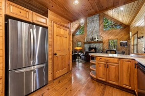 A kitchen with wooden cabinets, a stainless steel refrigerator, a stone fireplace, and a large window. - Peaceful Peaks - Mountain View | Hot Tub | Pet Friendly | Game Room | Firepit (Mineral Bluff)