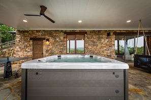 A hot tub in a stone-walled room with a ceiling fan, a door, and a view through a glass door. - Blue Mountain Retreat - Panoramic Mountain Views | Gourmet Kitchen | Expansive Decks | Dogs OK (Morganton)