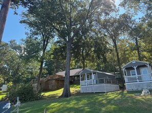 A cabin with a porch and a small deck, surrounded by trees and a grassy yard. - The Heron at The Woodlands at Water’s Edge (Many)