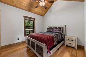 Ein Schlafzimmer mit Holzbalkendecke, einem Bett mit kariertem Überwurf, einem Nachttisch und einem Fenster mit Blick auf Bäume.