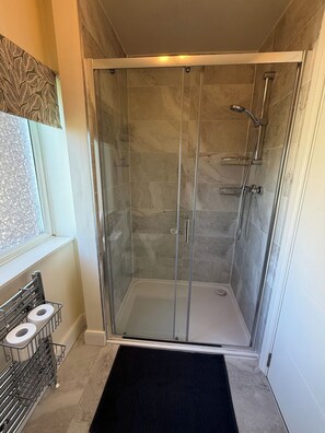 A glass-enclosed shower stall with a tiled floor, a metal towel rack with towels, and a window with patterned curtains. - Large Private Suite for Families & Workers Near Hospital (Greater London)
